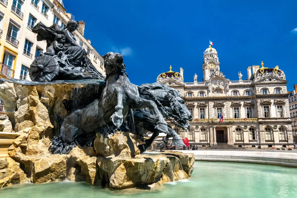 Masywna fontanna z dynamicznymi rzeźbami lwów i postaci mitologicznych, z historycznym budynkiem w tle.