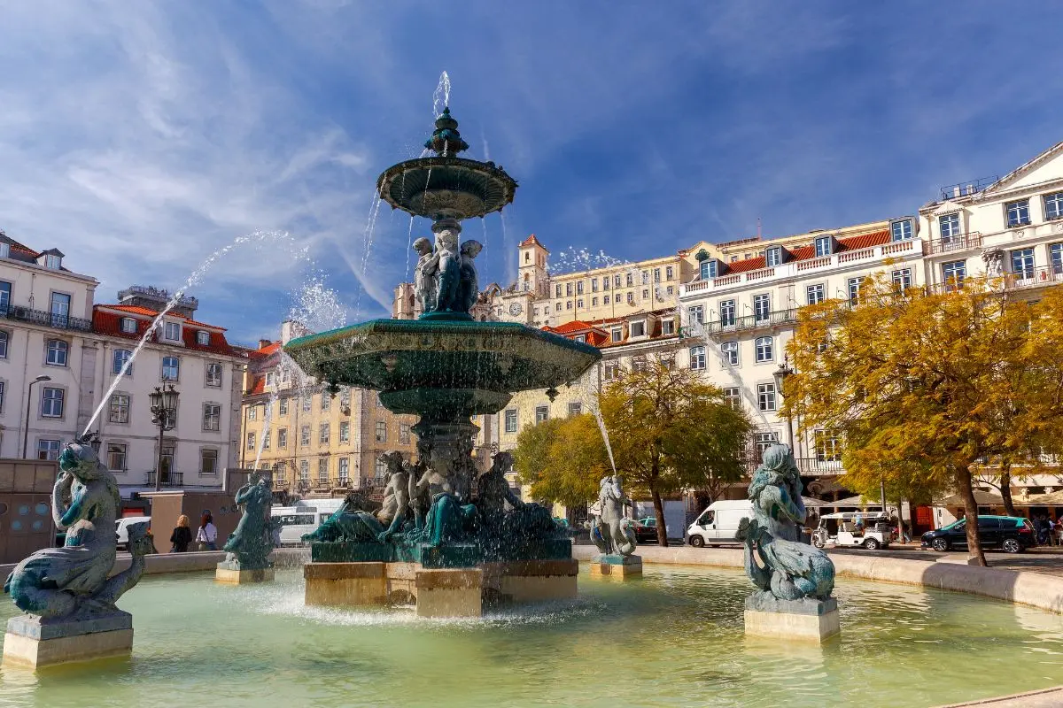 Klasyczna fontanna z posągami i strugami wody, otoczona miejskim zabudowaniem i żywym placem.