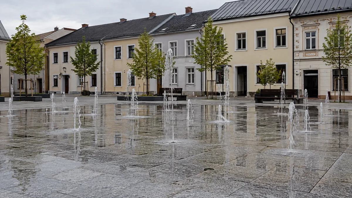 Fontanna posadzkowa z pionowymi strumieniami wody na brukowanym rynku małego miasta, w tle kamienice, popołudniowe światło